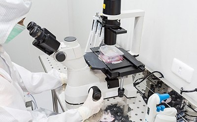 Scientist in full protective gown and mask looking through a microscope at cells in a T flask.