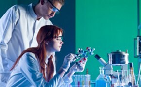 A laboratory setting with two individuals in white lab coats. One person is intently examining a molecular model, while the other observes from behind. Various laboratory equipment, including test tubes and a flask, is arranged on the table, with a green backdrop providing a vibrant contrast.