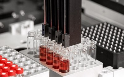 A close-up view of a laboratory automation system with multiple pipette tips positioned above several glass vials containing red liquid. 