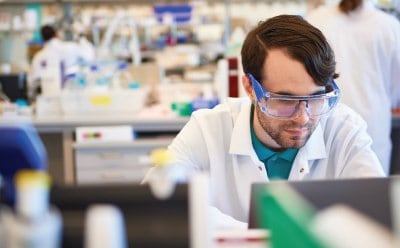 A person in a lab coat working in a well-equipped laboratory. Background includes various laboratory equipment and other individuals