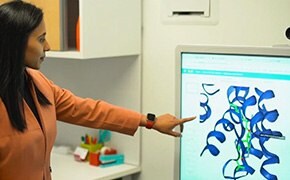 A woman in a peach blazer is demonstrating AI-powered AIDDISON™ software on a large touchscreen monitor. She is pointing at a colorful 3D molecular structure displayed on the screen, which represents molecular design, pharmacophore searches, and molecular docking used for effective drug discovery screening. The background shows an organized office environment with additional computer monitors and office supplies visible on a desk. The setting emphasizes the use of advanced technology in pharmaceutical research and development.