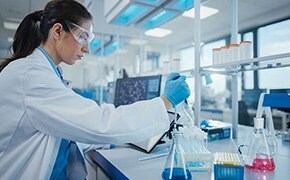 A scientist in a laboratory, wearing a white lab coat, safety glasses, and blue gloves, is working with analytical reagents, using a pipette to transfer a liquid into a flask, with various colorful solutions and lab equipment on the bench.