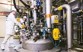 The image shows two technicians in a modern industrial laboratory working with large-scale chemical synthesis equipment. They are dressed in white lab coats and safety gear, indicating adherence to strict safety and quality control protocols. The setup includes a complex network of pipes, valves, and a large reaction vessel, illustrating advanced capabilities for custom chemical synthesis. This scene highlights the comprehensive services offered, including quality control, testing, documentation, bulk chemicals and contract manufacturing tailored to specific client needs.