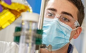 A scientist in a laboratory, wearing a white lab coat, safety glasses, a face mask, and purple gloves, is working with chemical biology tools. He is carefully pouring a yellow liquid from a beaker into a specialized glass apparatus. The background suggests a well-equipped lab environment focused on chemical biology research.