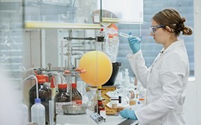 A female scientist in a lab coat and safety goggles is working in a laboratory, holding a pipette and carefully adding a substance to a reaction vessel. The lab bench is filled with various bottles and containers of chemical reagents, along with glassware and equipment used in chemical synthesis. The image portrays the precise and methodical work involved in handling chemical building blocks, essential for developing new compounds and conducting advanced research.