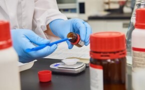 A scientist in a laboratory, wearing blue gloves, is handling a brown bottle of SigmaAldrich™ chemical reagents, with various other bottles and lab equipment visible on the bench, indicating an active research setting.