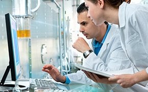 Two scientists in a modern laboratory, wearing white lab coats, are using ChemisTwin™ digital reference materials for conducting qualitative NMR analysis and quantification of compounds. They are intently looking at a computer screen, surrounded by advanced lab equipment and glassware, discussing their findings.