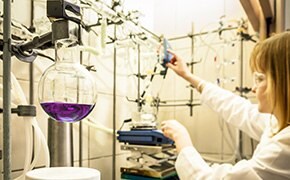 A scientist in a laboratory setting, wearing a white lab coat and safety goggles, is working with click chemistry reagents. She is adjusting a piece of equipment connected to a complex glassware setup, including a round-bottom flask containing a purple solution. The lab is equipped with various tubes and connectors essential for chemical reactions.