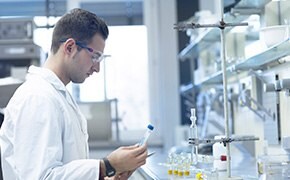 A scientist wearing a white lab coat and safety glasses is working in a laboratory. He is holding a test tube with a blue cap in one hand and a small device in the other. The laboratory bench is equipped with various glassware, including a vertical glass apparatus held by a clamp. The background shows shelves with additional laboratory equipment. The image is associated with the use of DyNAbind® DNA encoded library kits and DEL data software, which are tools for fragment-based and high-throughput screening in drug discovery.