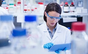 A female scientist wearing safety goggles and blue gloves is working in a laboratory, holding a tablet. She is surrounded by various brown glass lab bottles and white plastic containers with red caps, which contain SigmaAldrich™ chemicals. The image highlights the range of chemicals provided by SigmaAldrich™, including solvents, acids, salts, caustic alkalis and bases, metals, and test kits. The lab setting underscores the professional and precise nature of the work carried out using these high-quality chemical products.