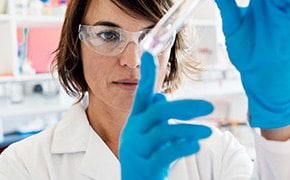 A scientist in a laboratory, wearing a white lab coat, safety glasses, and blue gloves, is closely examining a test tube containing a pink solution, indicative of research involving polymeric microspheres and nanoparticles.