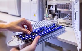 A tray of blue-capped vials are placed into a laboratory instrument, likely a chromatograph, for chemical analysis. The machine is partially open, revealing its internal components, indicating preparation for testing or calibration in a chemistry lab setting.