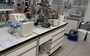 The image shows a laboratory setup featuring the SYNPLE 2 automated synthesizer, prominently displayed on the left side with its digital screen active. Surrounding the synthesizer are various pieces of laboratory equipment used in chemical synthesis, including reaction vessels, bottles of reagents, and analytical instruments. The lab bench is organized, with detailed notes and safety protocols visible, indicating a professional environment dedicated to process chemistry.