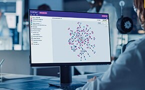 A scientist is sitting at a desk, using a computer with the SYNTHIA™ retrosynthesis software displayed on the monitor. The screen shows a complex network of colorful nodes and connections representing synthetic routes to complex molecules. The software interface includes options for filtering and analysis. The laboratory environment is modern, with other scientists working in the background, emphasizing the collaborative and high-tech nature of the research.