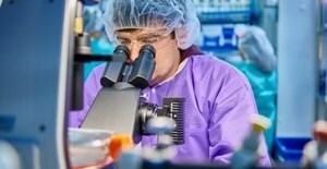 Scientist wears PPE (head net + lab coat) and peers into microscope.