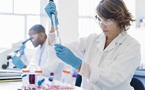 Female scientist wearing white lab coat, blue nitrile gloves, and safety goggles pipetting a protein solution into a centrifugal tube. Male scientist wearing same protection clothing looking into microscope is visible in the background.