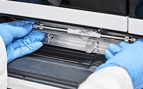 HPLC column is installed in lab instrument. The hands of a lab worker are shown wearing blue nitrile gloves.