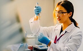 Dark-haired female scientist wearing in blue nitrile gloves, white lab coat, and safety goggles pipettes solution into a centrifugal tube