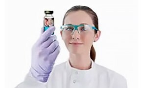 A female scientist wearing protective goggles holds up a sterile drug product.