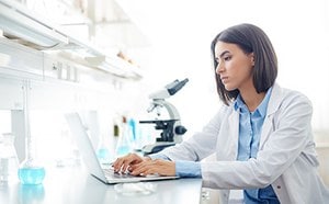 A laboratory setting with a person wearing a white lab coat, sitting at a desk and typing on a laptop. In the background, there is a microscope and various laboratory glassware containing colorful liquids. The environment is bright and well-lit, suggesting a professional scientific workspace.