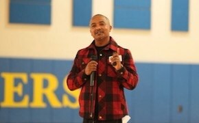 Abdi Wege wears a red flannel and holds a microphone in his right hand.