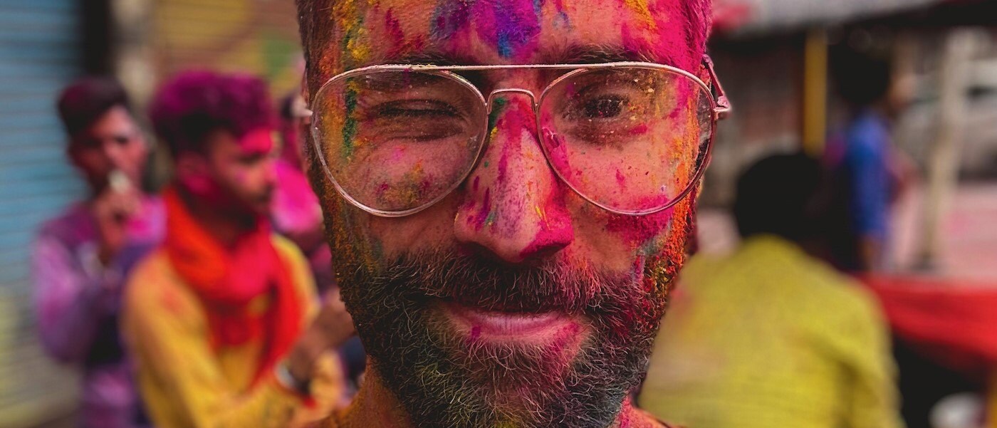 A close-up of André Silva’s face. Silva wears glasses and squints one eye. A number of colors appear on his face, evidence that he participated in a Holi celebration.