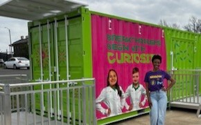 Danielle Chavis wears a SPARK t-shirt and stands in front of the Curiosity Cube which is bright green, has images of two young scientists with the words “Breakthroughs Begin with Curiosity” on the front. 