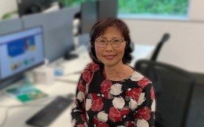 Grace Yee wears a headset and sits at her desk. In the background is her desktop computer. 