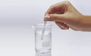 A hand is holding a test strip above a beaker filled with clear liquid. The beaker is labeled with a measurement of 100 ml and is placed on a light background. The test strip is being lowered into the water.