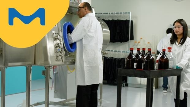 A male scientist in a lab coat is using an isolator, while a colleague female scientist in a lab coat is transferring chemicals into the training facility behind him.